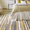 Hand-tufted rug with vibrant stripes of yellow, brown, black, and off-white, placed beside a cozy bed.